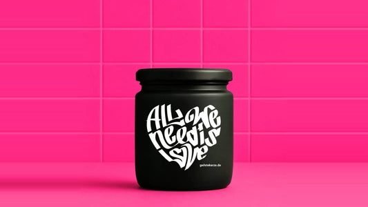 Duftkerze im Glas mit dem Aufdruck ALL WE NEED IS LOVE, vor einer pinken Fliesenwand